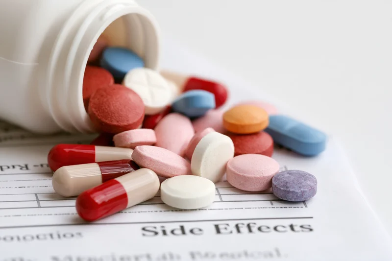 Assorted prescription pills and capsules spilling from a bottle onto a document labeled side effects