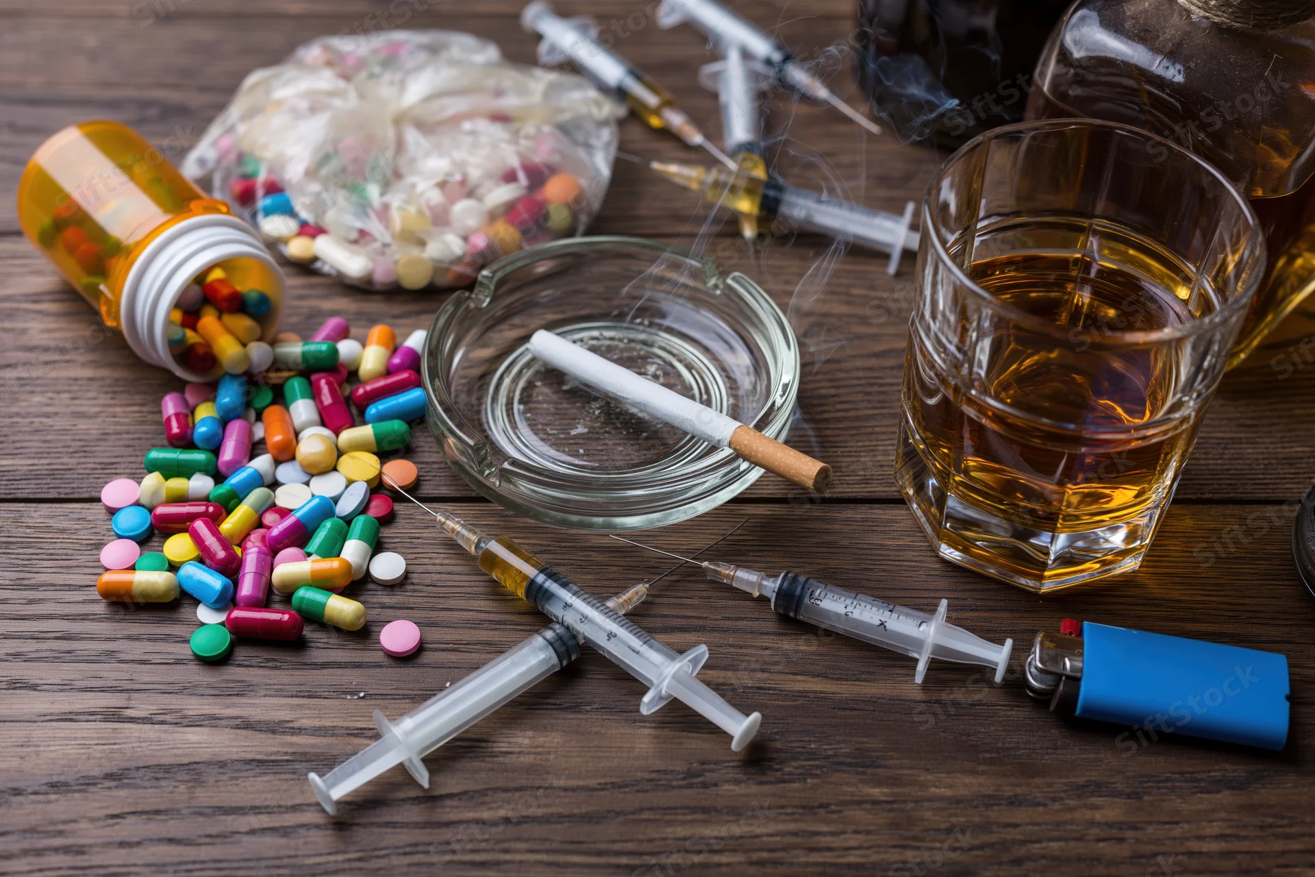 Assorted substance abuse paraphernalia including syringes, pills, alcohol, and cigarettes arranged on a dark wooden table