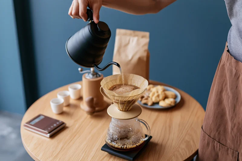 Barista pouring hot water from a gooseneck kettle into a paper filter over a glass carafe on a wooden table