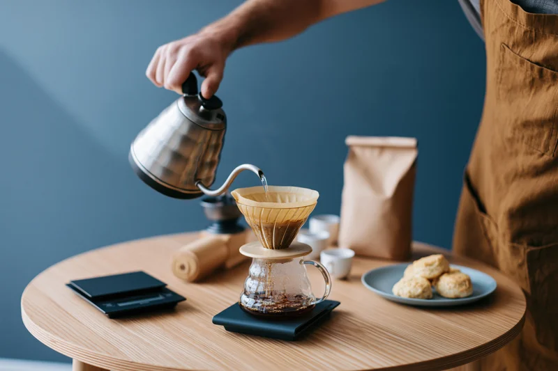 Barista pouring hot water from a gooseneck kettle into a paper filter over a glass carafe on a wooden table