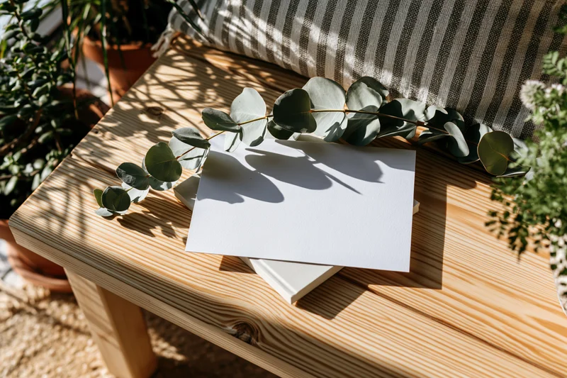 Blank white card resting on wooden table with eucalyptus branch and striped cushion nearby