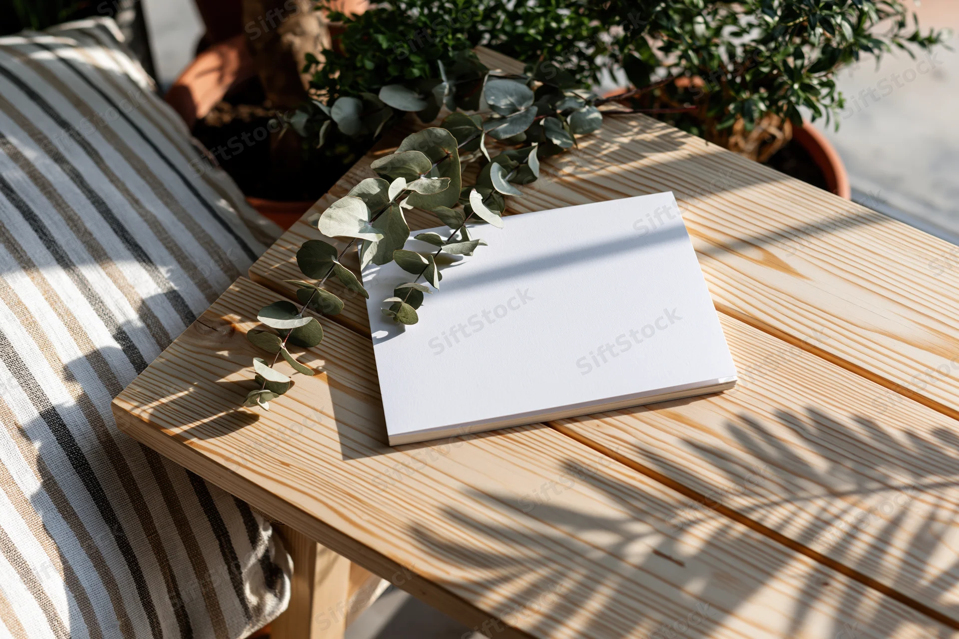 Blank white card lying flat on wooden table with eucalyptus branch and striped cushion beside it outdoors