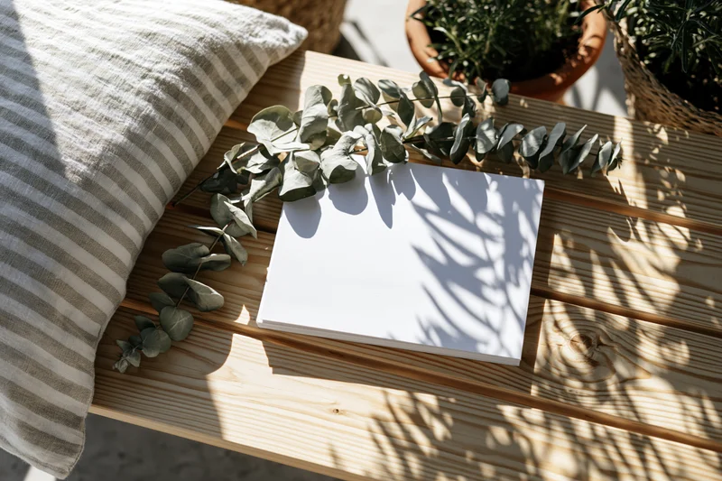 Blank white card lying flat on light wooden bench with eucalyptus branches and striped pillow beside it