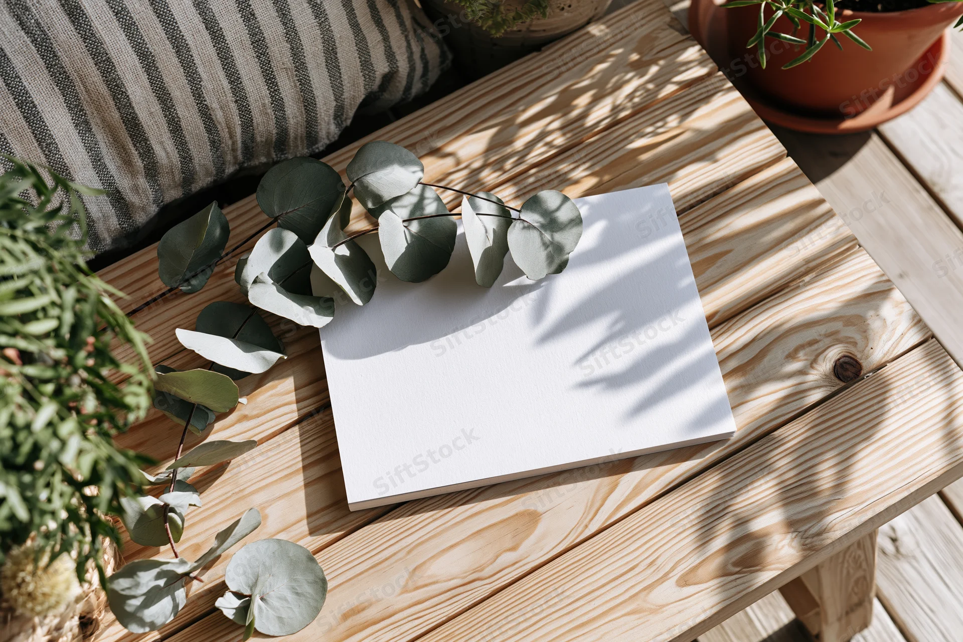 Blank white card lying flat on light wooden table with eucalyptus leaves and potted plants nearby