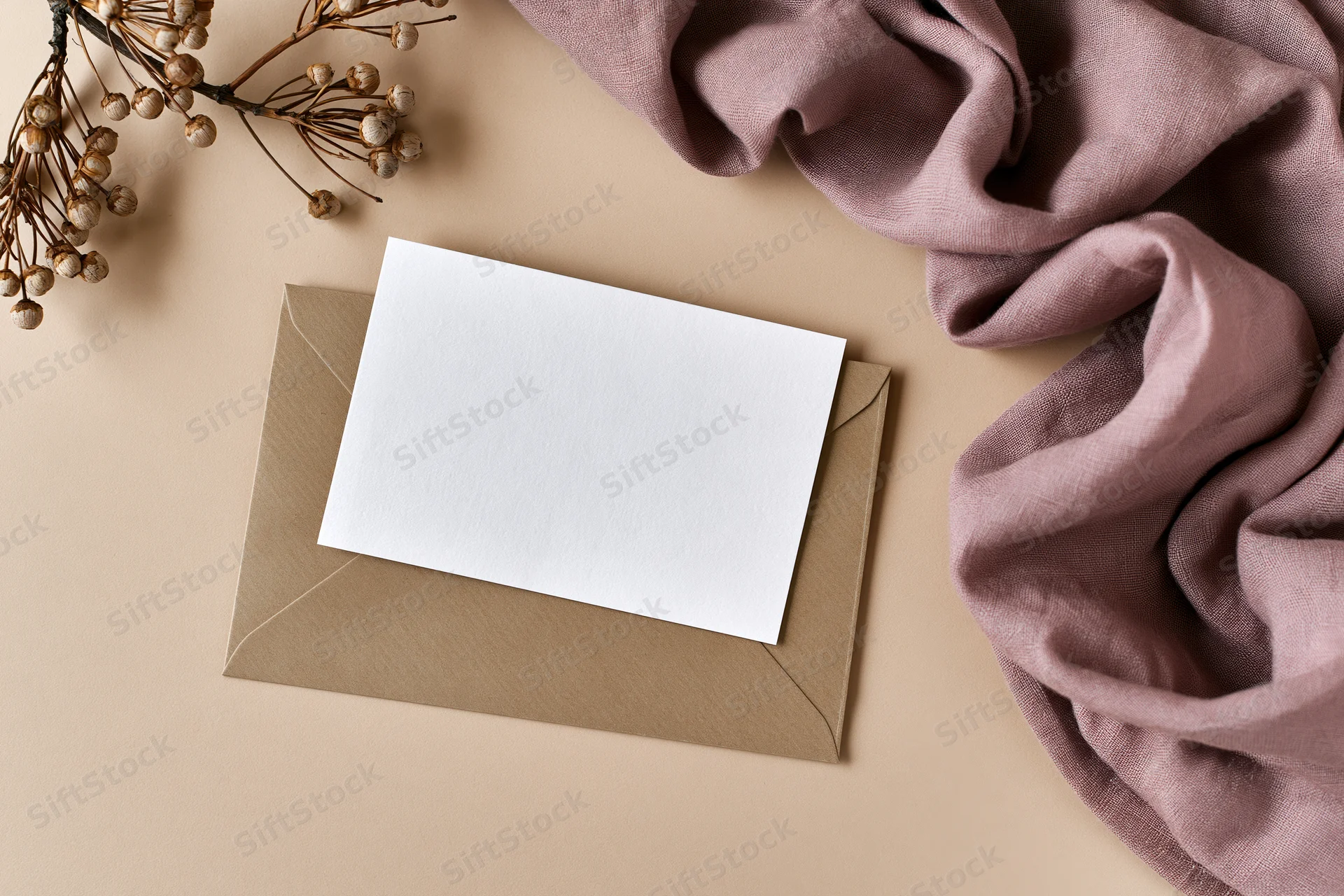 Blank white card on kraft envelope with dried flowers and mauve fabric in flat lay arrangement