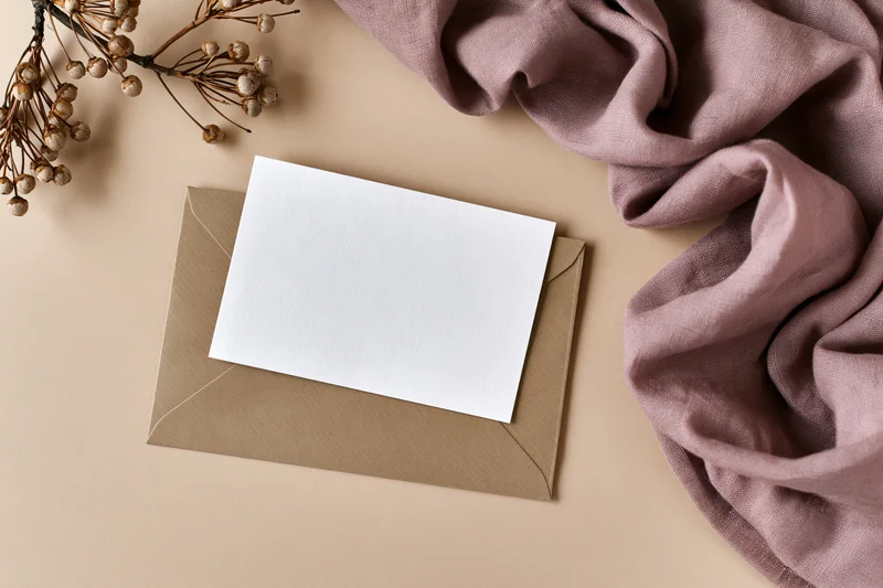 Blank white card on kraft envelope with dried flowers and mauve fabric in flat lay arrangement
