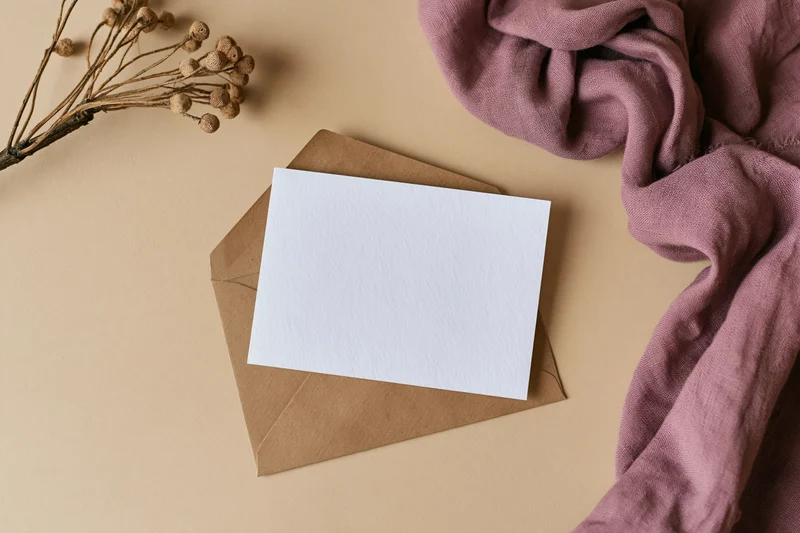 Blank white card on kraft envelope with dried flowers and mauve fabric in flat lay arrangement