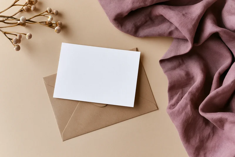 Blank white card on kraft envelope with dried flowers and mauve fabric in flat lay arrangement