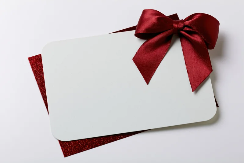 Blank white gift card with red satin bow on glittery red base, isolated on white background