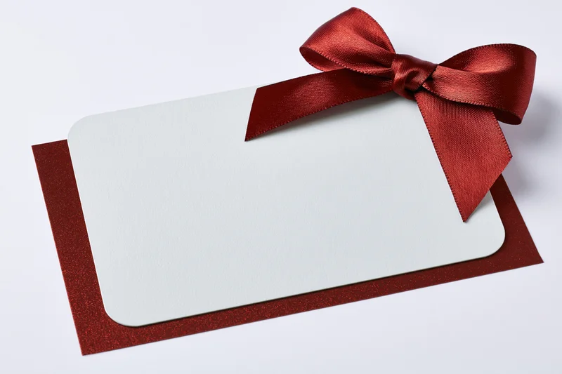 Blank white gift card with red ribbon bow on glittery red envelope against white background