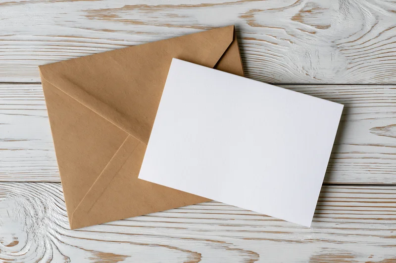 Blank white greeting card resting on a kraft paper envelope on a weathered white wooden surface