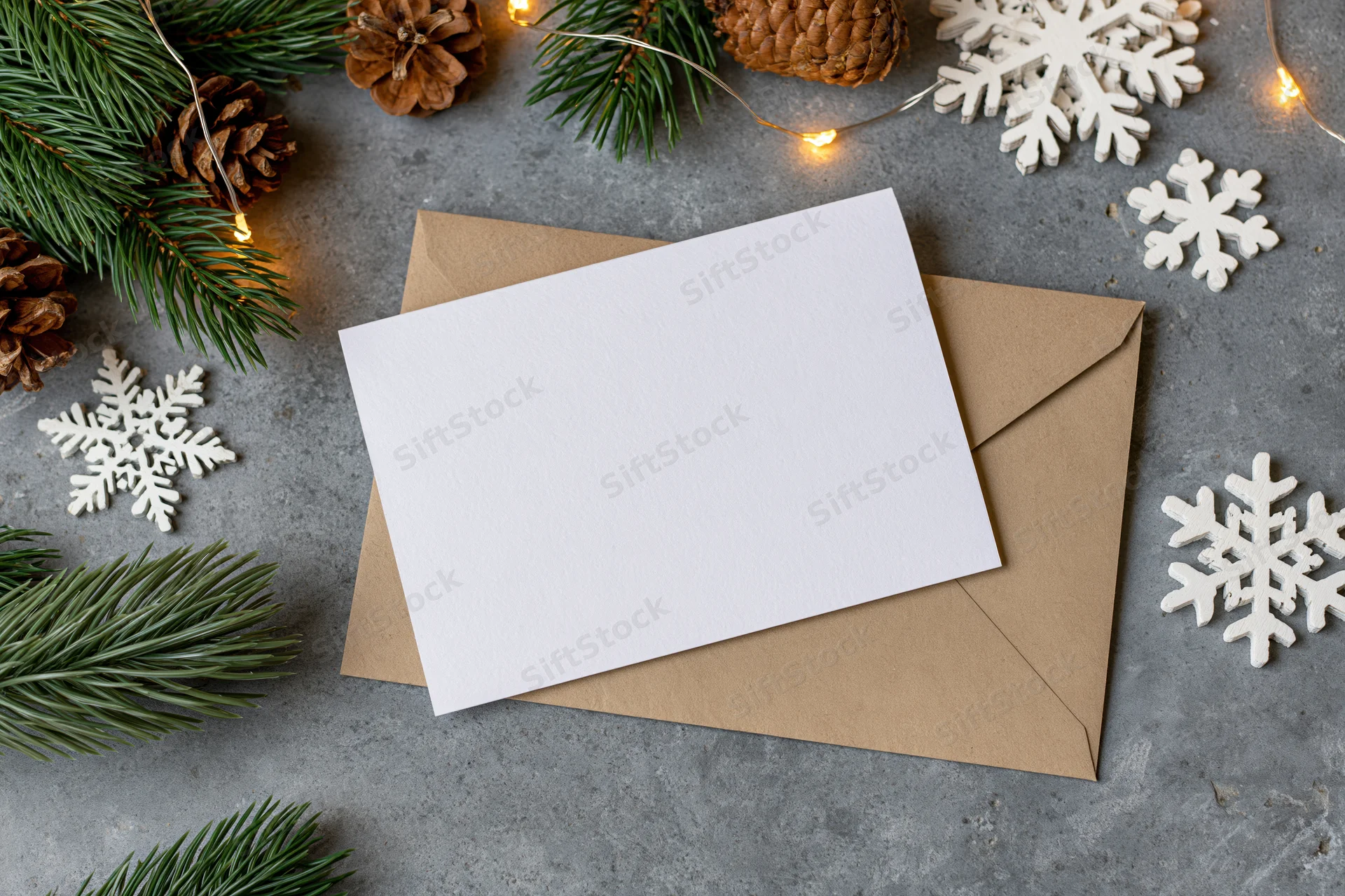 Blank white greeting card on kraft envelope surrounded by pine branches, pinecones, snowflakes and string lights on gray surface