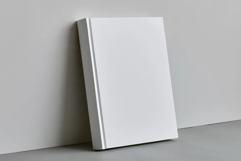 Blank white hardcover book leaning against a light gray wall in a minimalist studio setting