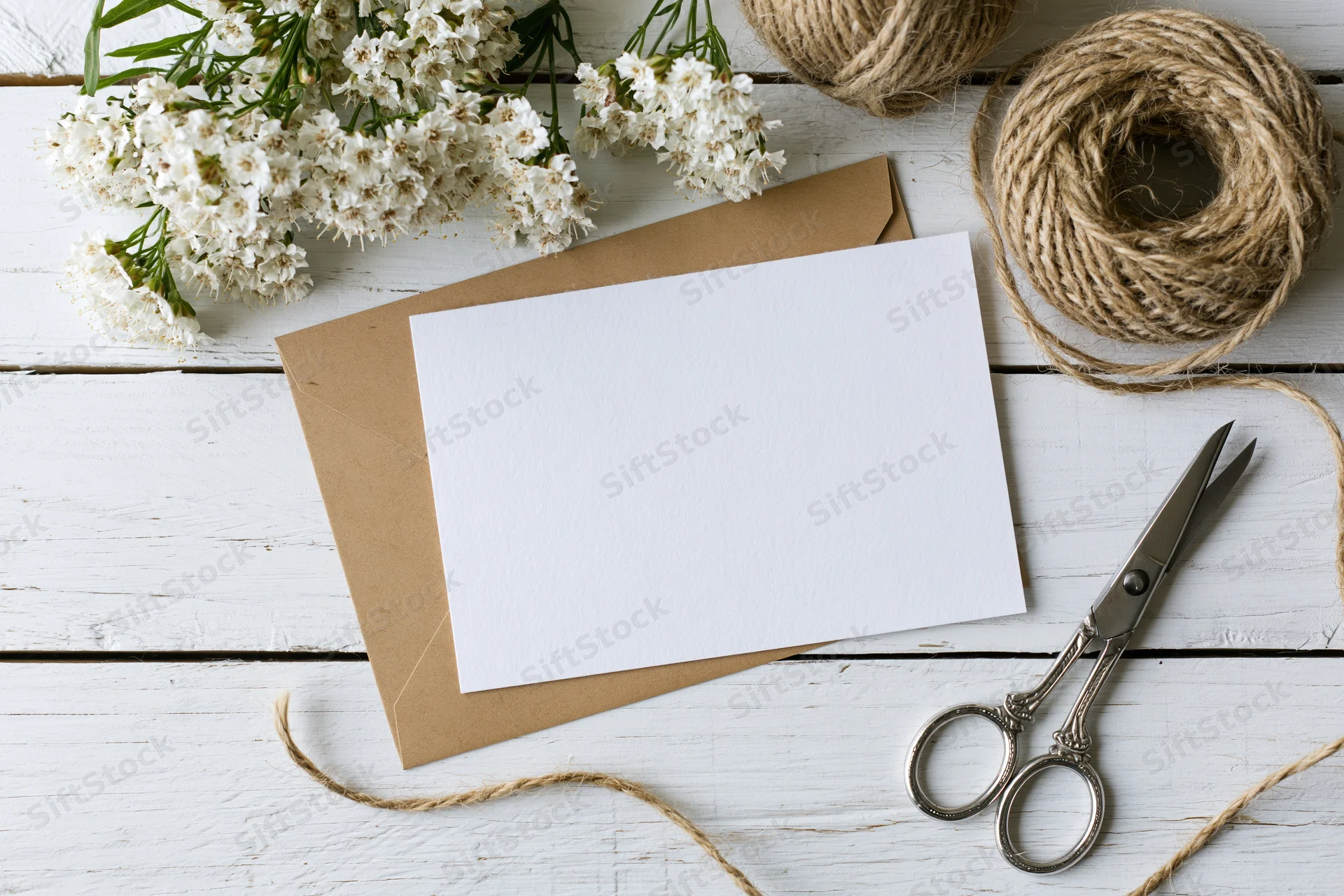 Blank white notecard on kraft envelope with white flowers, twine and scissors on whitewashed wood surface