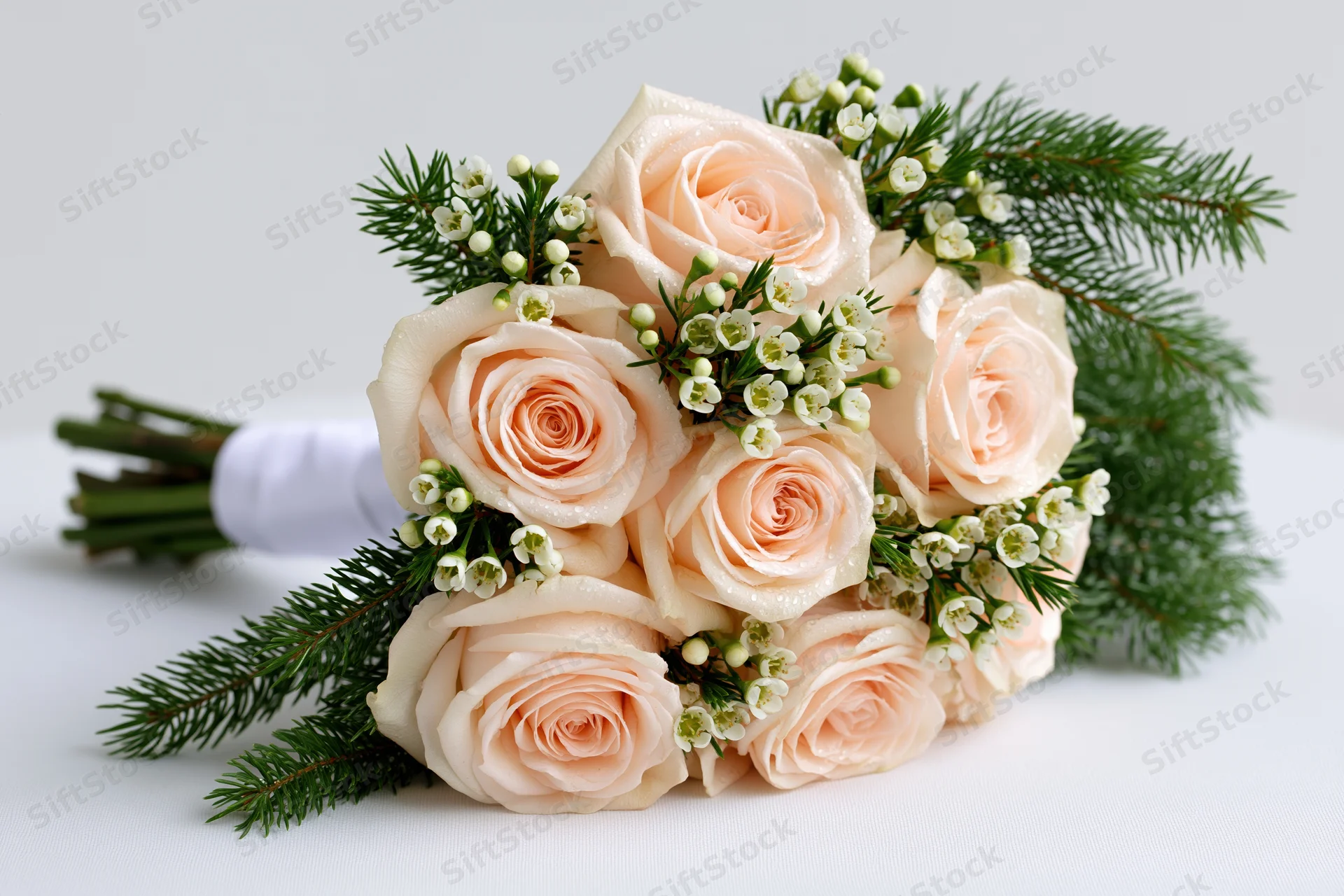 Bridal bouquet of pale blush roses and small white wax flowers with evergreen sprigs on white surface