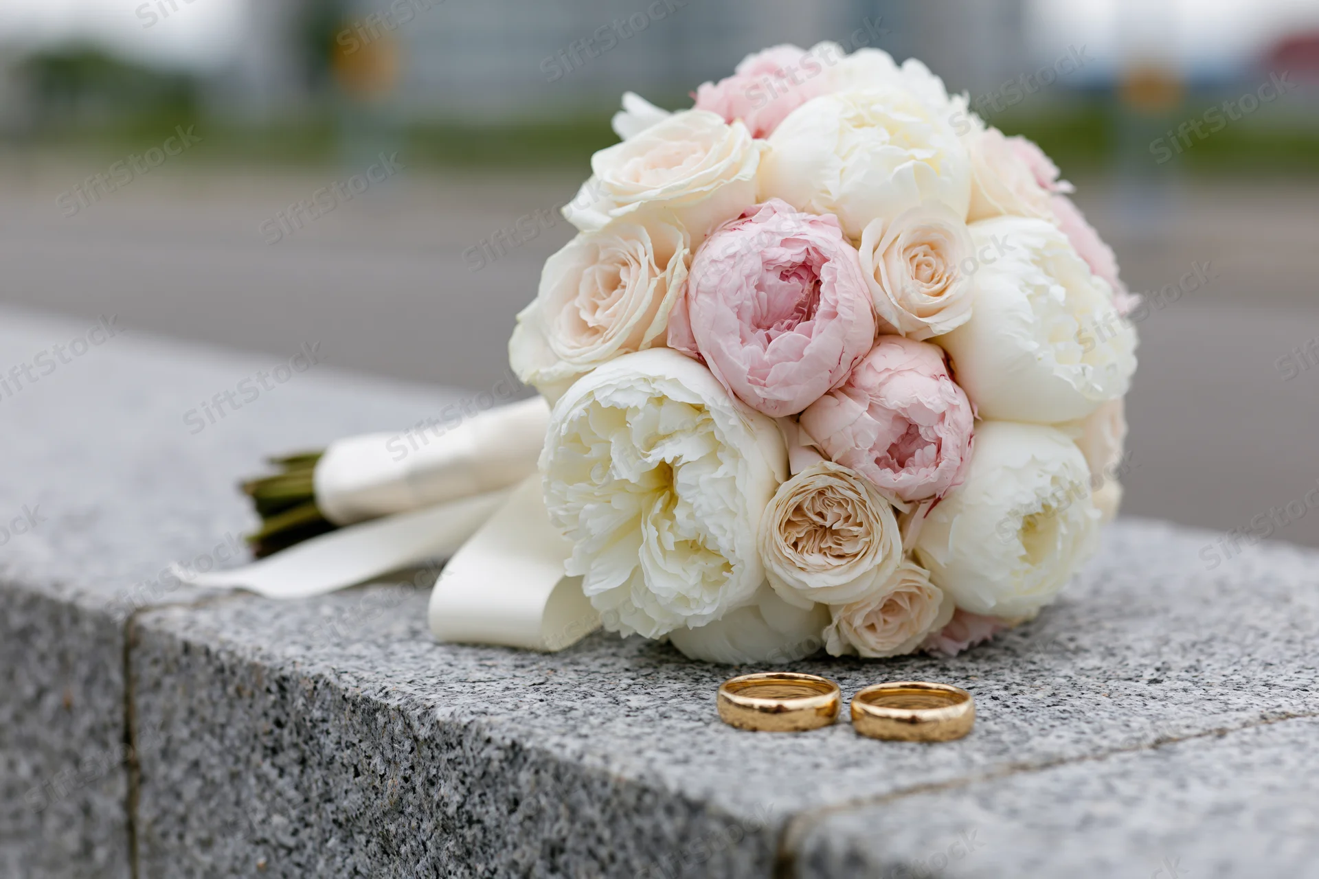 Bridal bouquet of white and blush peonies with gold wedding rings on a granite ledge outdoors