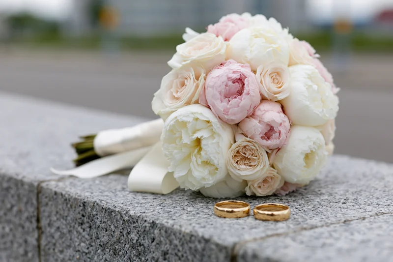 Bridal bouquet of white and blush peonies with gold wedding rings on a granite ledge outdoors