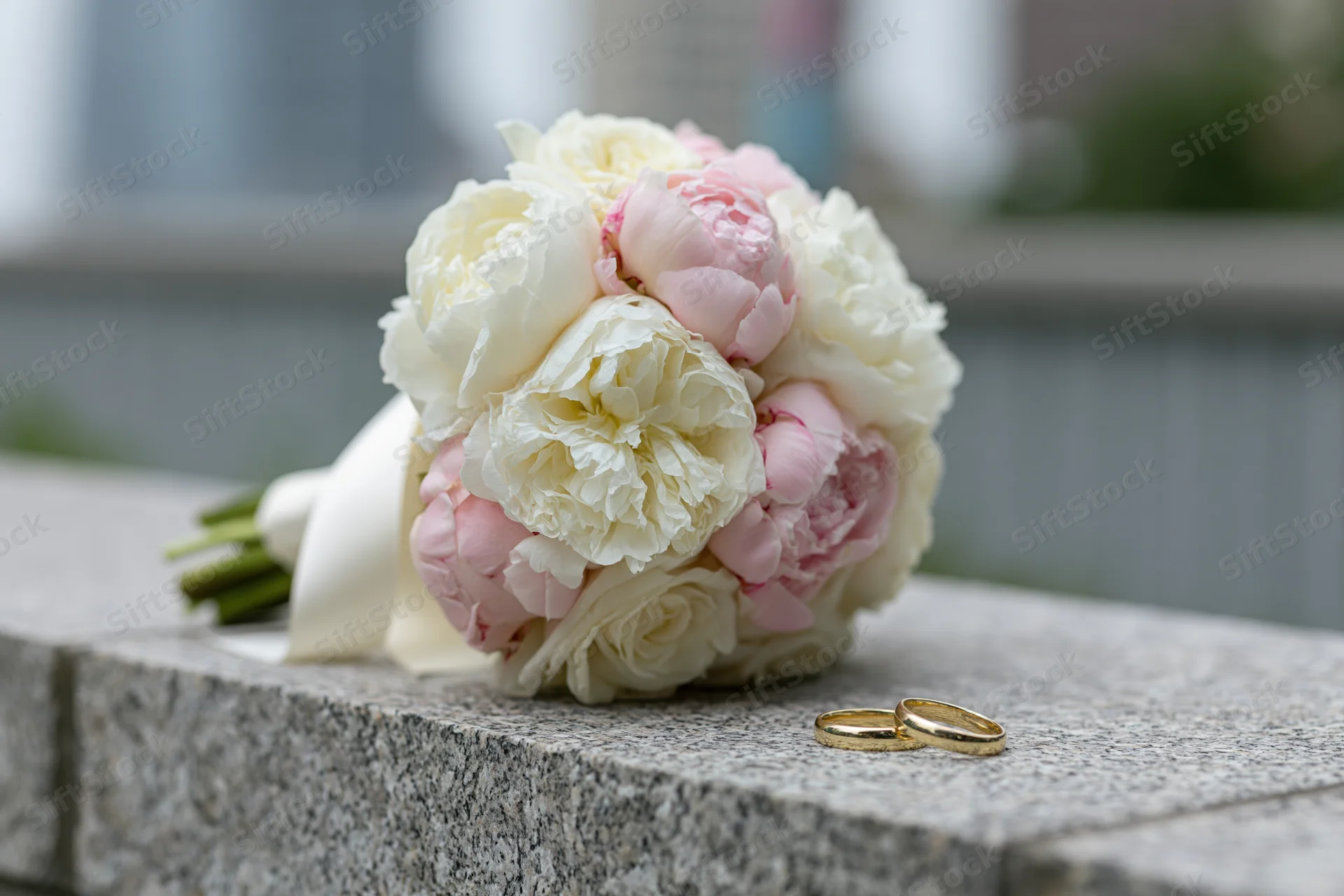 Bridal bouquet of white and blush peonies resting on a stone ledge with two gold wedding rings beside it