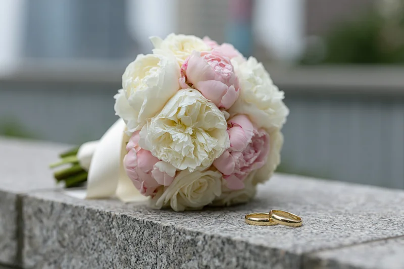 Bridal bouquet of white and blush peonies resting on a stone ledge with two gold wedding rings beside it
