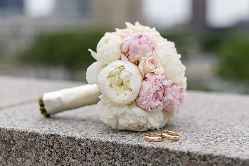 Bridal bouquet of white and blush peonies resting on a granite surface with two gold wedding rings beside it
