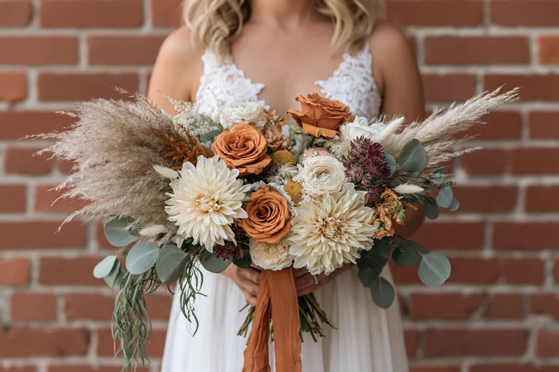 Bride holding large cascading bouquet of orange and white flowers with pampas grass and eucalyptus against brick wall