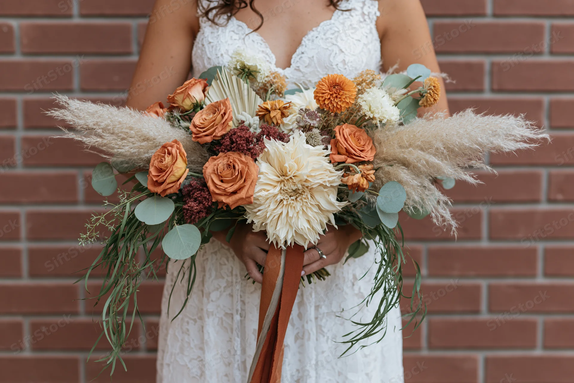 Bride holding large cascading bouquet of orange roses and pampas grass against brick wall