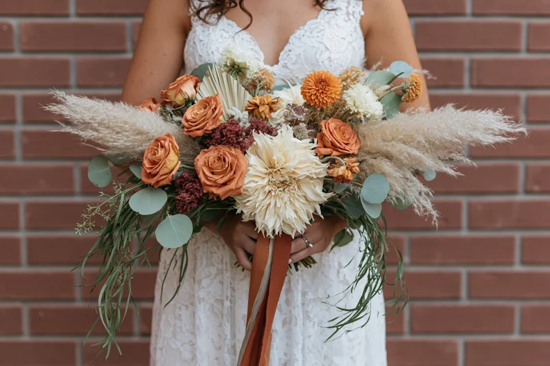 Bride holding large cascading bouquet of orange roses and pampas grass against brick wall