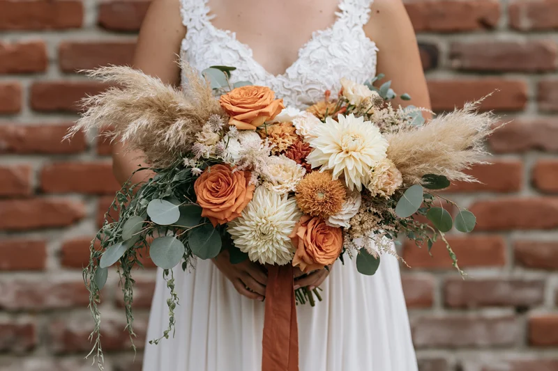 Bride holding a large cascading bouquet of orange roses and white dahlias with pampas grass and eucalyptus against a brick wall