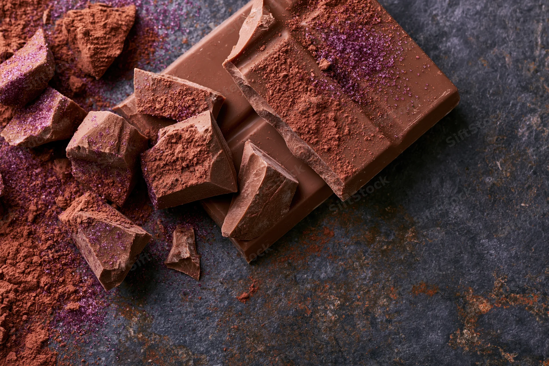Broken milk chocolate bar pieces and scattered chunks dusted with cocoa powder on a dark textured surface