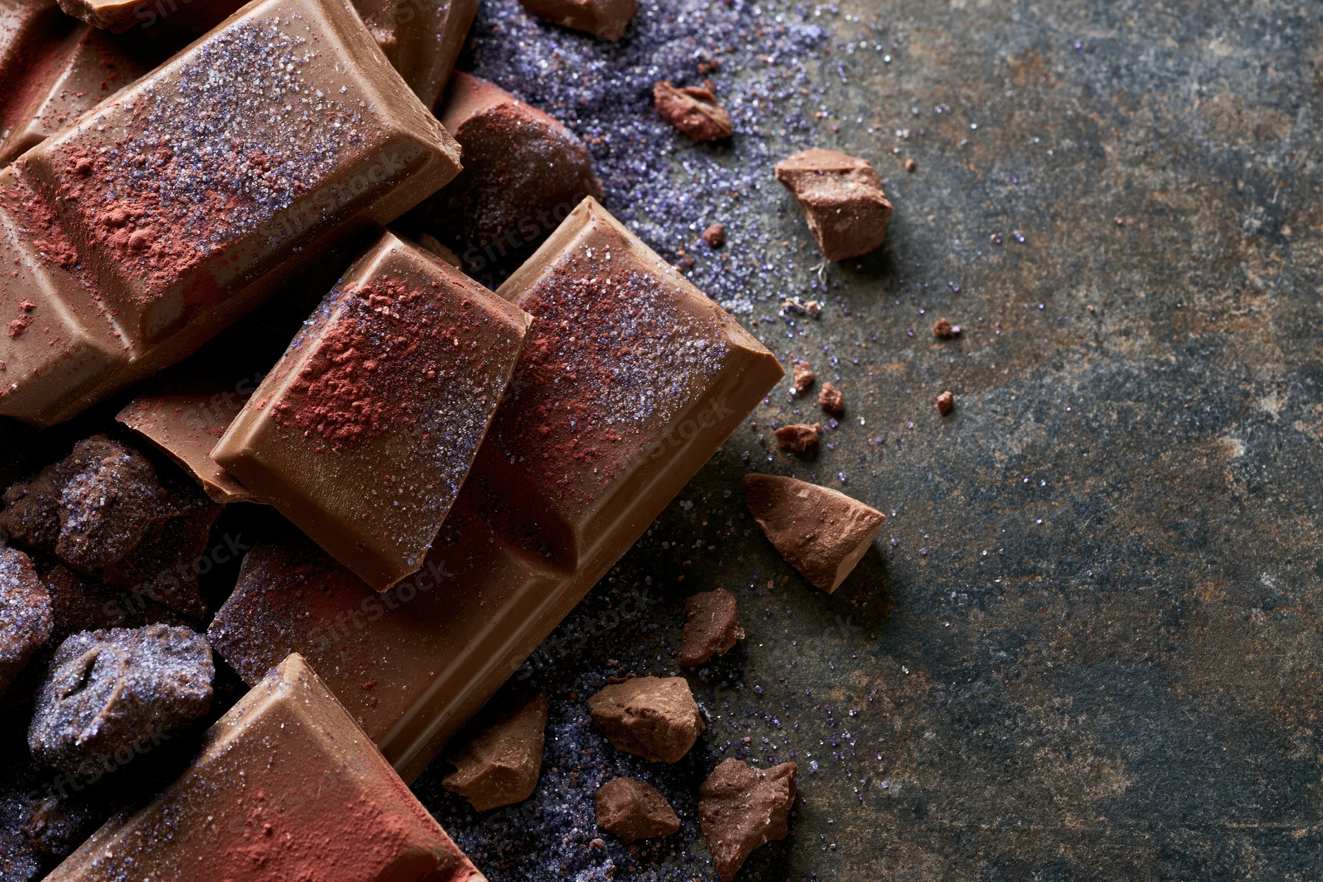 Broken milk chocolate bar pieces and scattered chunks dusted with cocoa powder on a dark textured surface