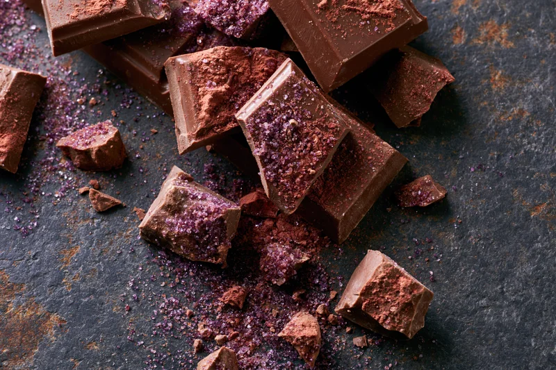 Broken milk chocolate pieces scattered on a dark rustic surface dusted with purple glitter and cocoa powder