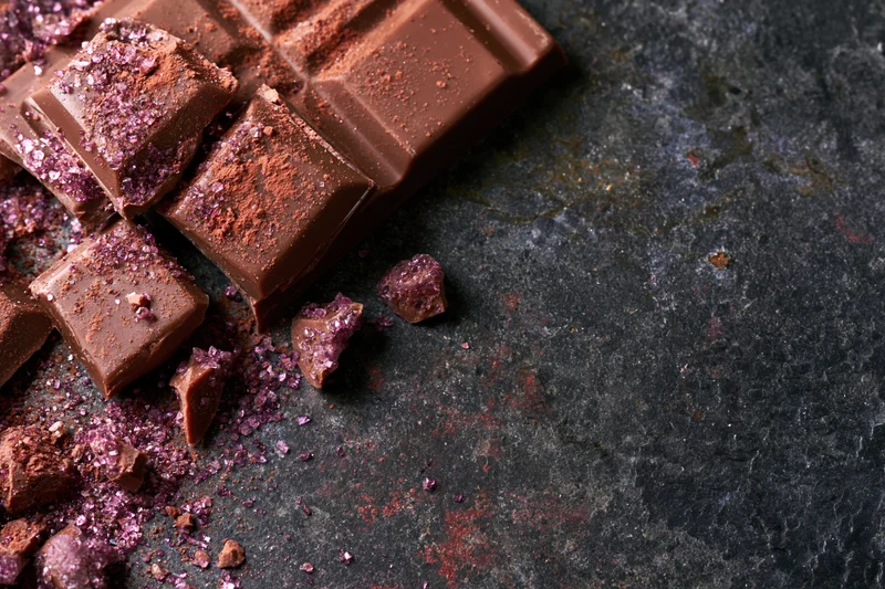 Broken milk chocolate bar pieces and scattered chunks dusted with purple cocoa powder on dark stone surface