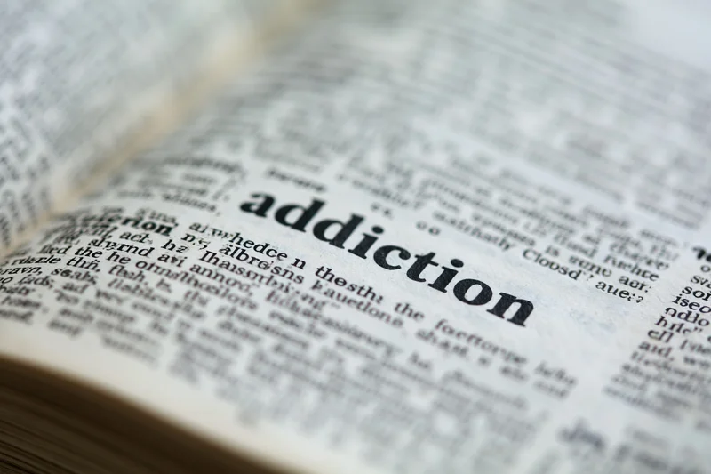 Open dictionary page showing the word addiction in bold print with blurred surrounding text