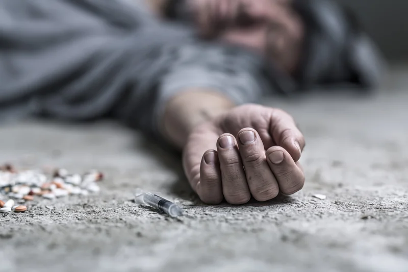 Pale hand lying limp on grimy concrete beside syringe and scattered pills