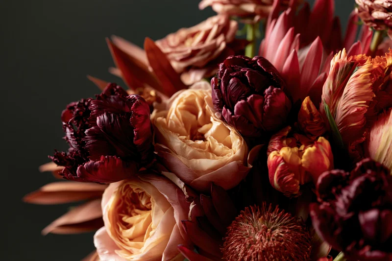 Close-up of a richly layered autumn floral arrangement featuring roses, tulips and peonies in burgundy, peach and cream tones against a dark background