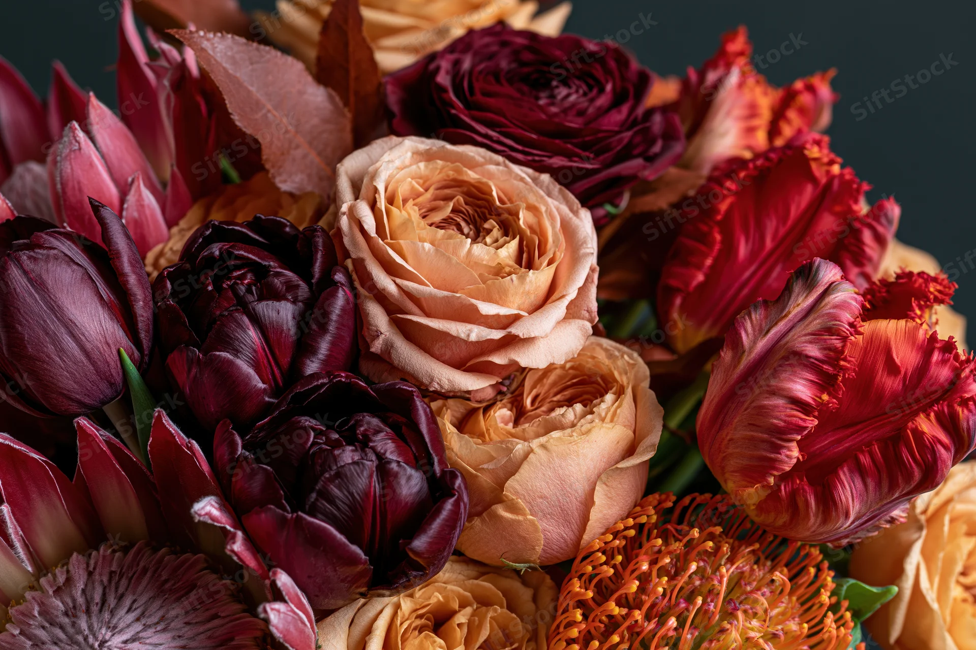 Close-up of a richly layered autumn floral arrangement featuring roses and tulips in deep burgundy, peach, and orange hues