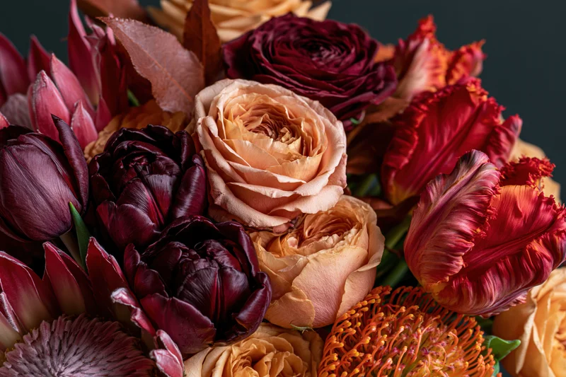 Close-up of a richly layered autumn floral arrangement featuring roses and tulips in deep burgundy, peach, and orange hues