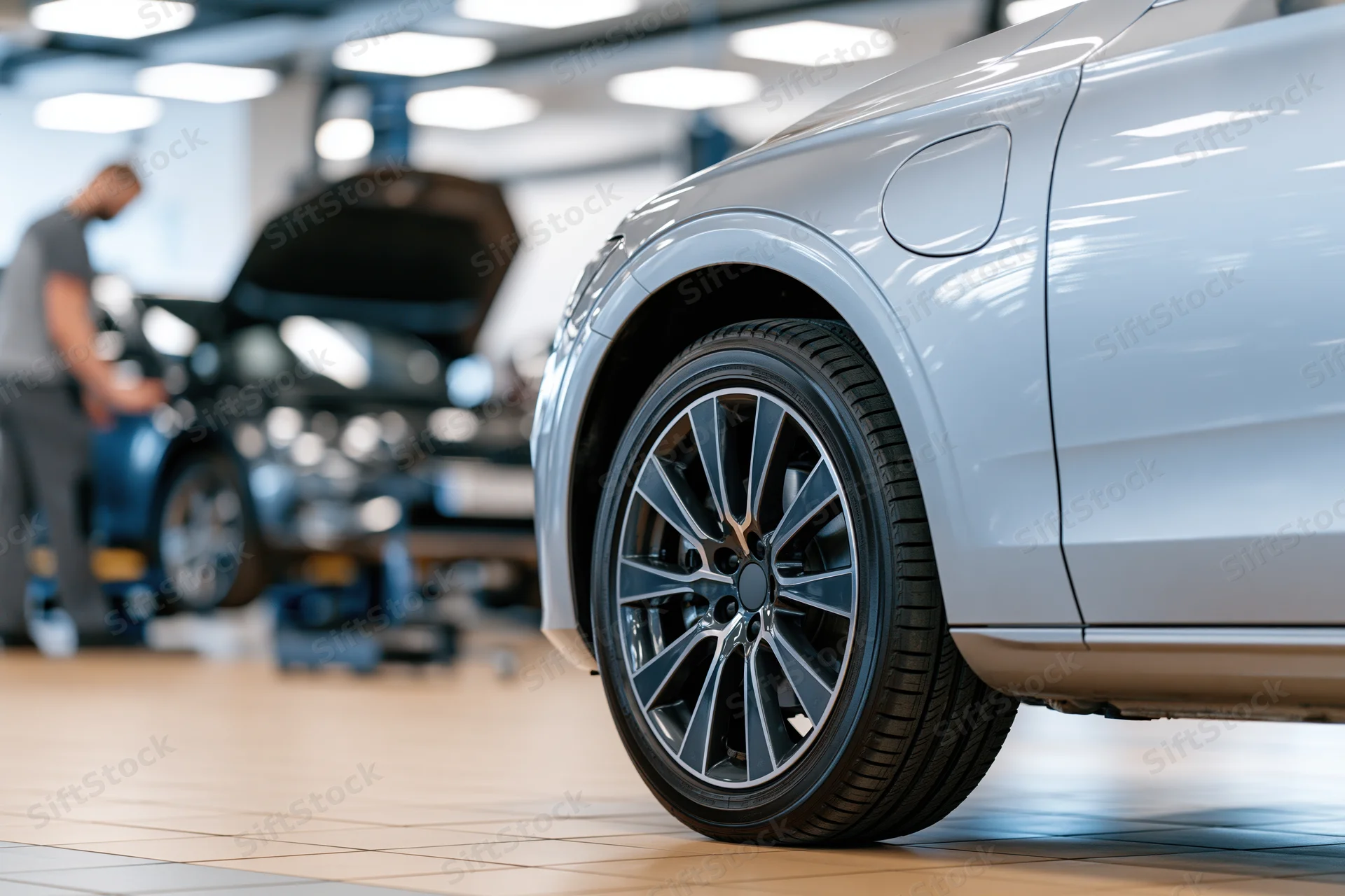 Close-up of a silver luxury sedan's front wheel and alloy rim in a garage with a mechanic working on another car in the background