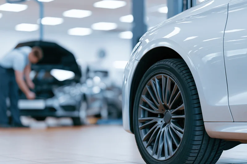 Close-up of a silver luxury sedan's front wheel and rim in a garage with a mechanic working on another car in the background
