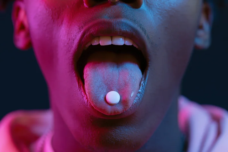 Young person with open mouth and tongue extended holding a white pill under neon pink and blue light