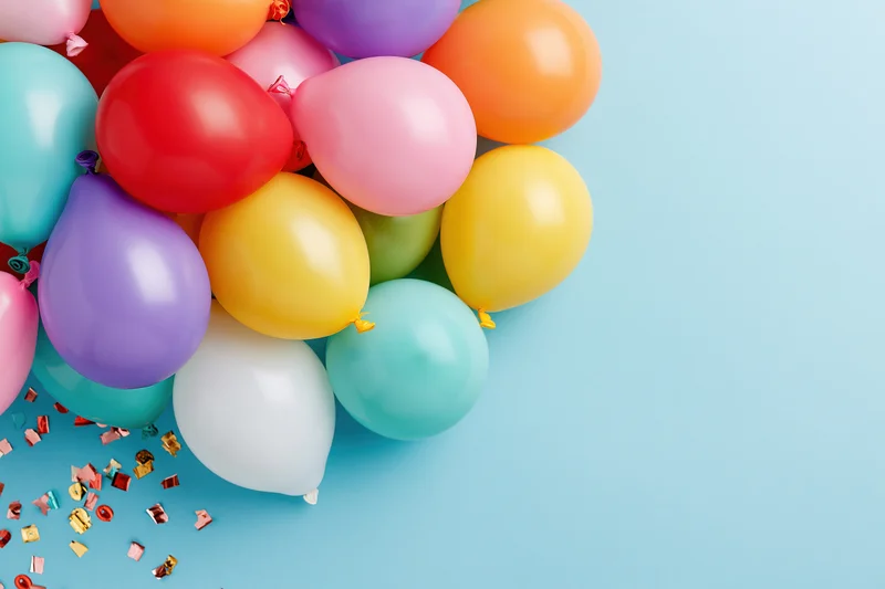 Cluster of multicolored latex balloons with confetti on blue background, left side arrangement with copyspace