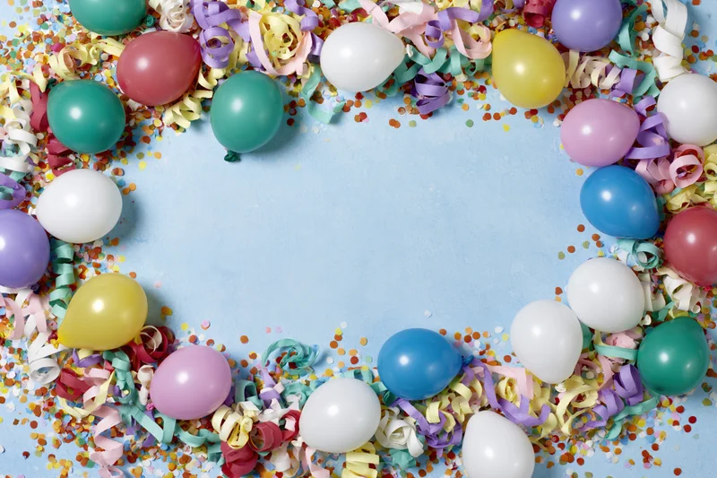 Colorful party balloons and curled paper streamers arranged in a circular frame on a blue surface with confetti and copyspace