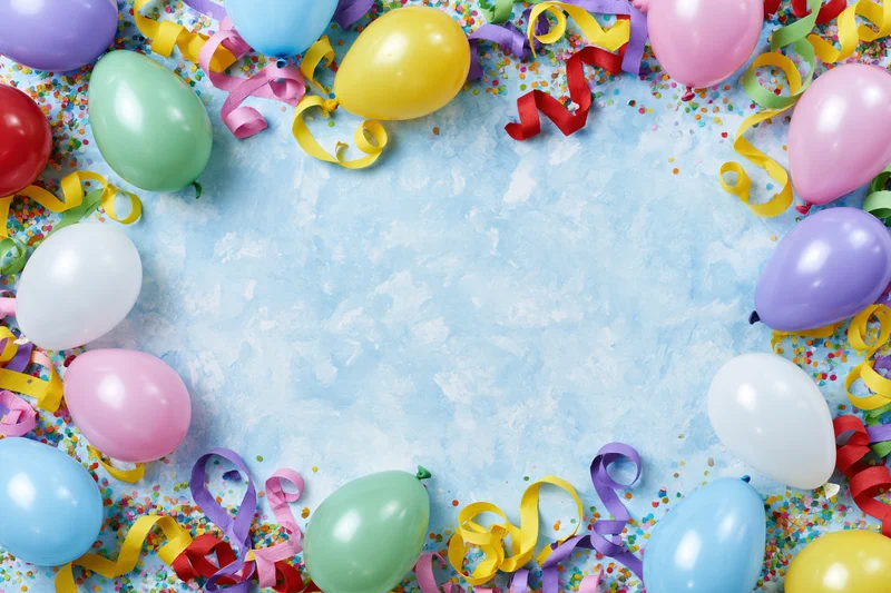Colorful party balloons and curled paper streamers arranged in a border around a blue textured surface with confetti, leaving central copyspace