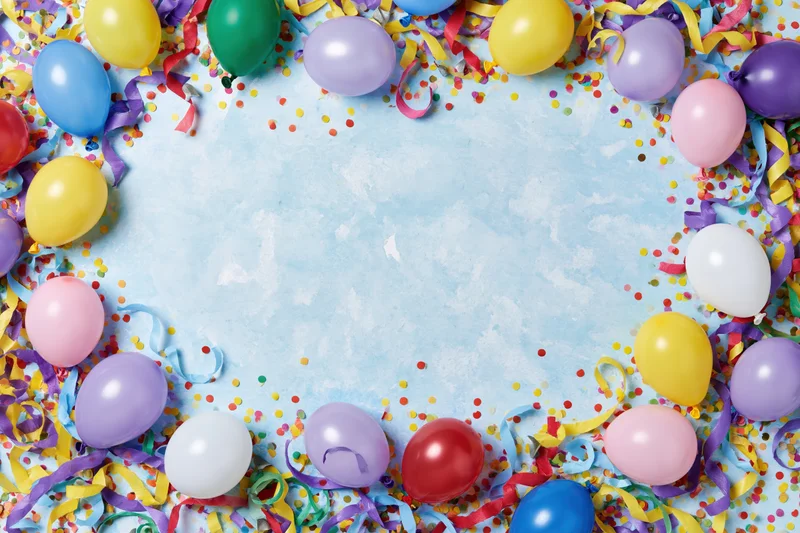 Colorful party balloons and streamers arranged in a circular frame around a light blue textured background with confetti