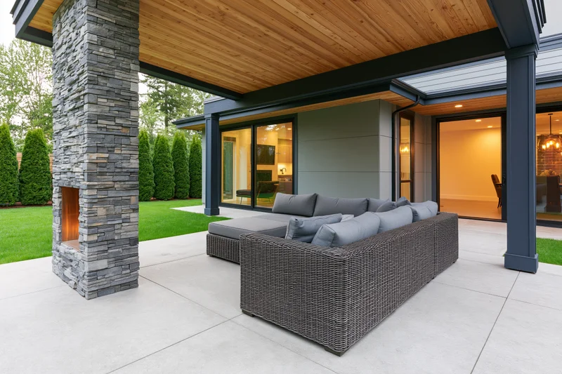 Modern covered patio with stacked-stone fireplace and wicker sectional sofa on concrete flooring