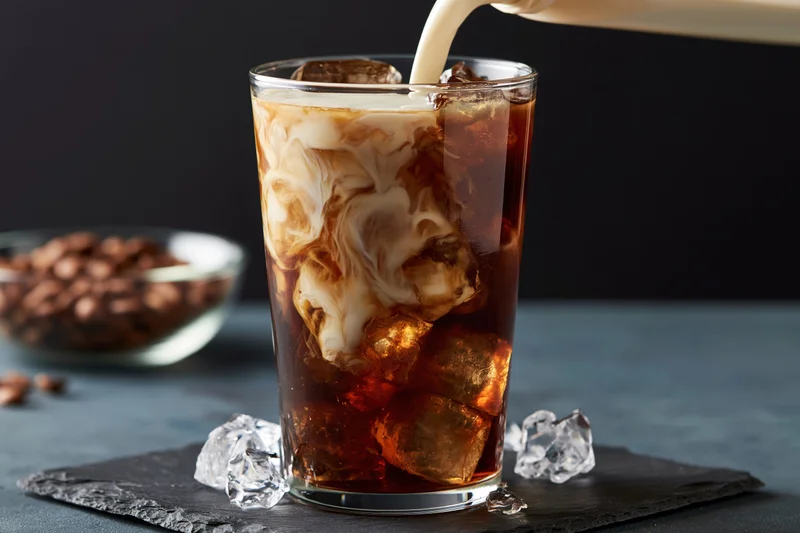 Cream being poured into a tall glass of iced coffee with ice cubes on a slate tray
