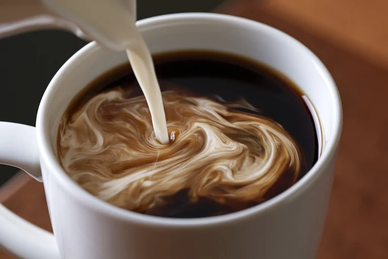 Cream being poured into a white ceramic mug of black coffee, creating swirling patterns on the surface