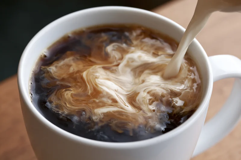 Cream being poured into a white ceramic mug of black coffee on a wooden surface