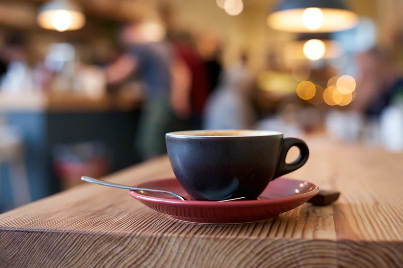 Dark ceramic espresso cup with spoon on red saucer on wooden table in cafe interior