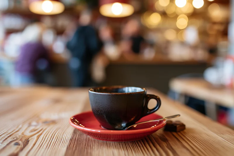 Dark ceramic espresso cup with spoon on red saucer on wooden table in cafe interior
